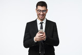 Indoor portrait of young European Caucasian male in formal clothes and eyeglasses isolated on gray background holding cellphone in front of him, typing or browsing with happy face and open smile