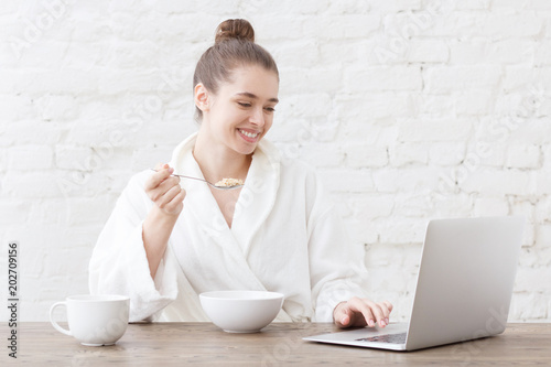 Studio portrait of young good-looking European Caucasian girl wearing white bathrobe in morning using laptop during breakfast checking daily emails and private messages showing happy friendly smile photo