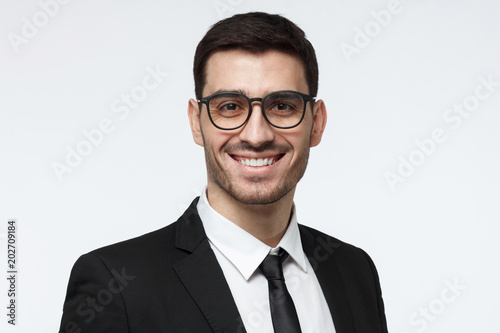 Horizontal headshot of young European Caucasian man in formal clothes isolated on gray background looking through glasses and smiling positively, feeling confident about success in his business