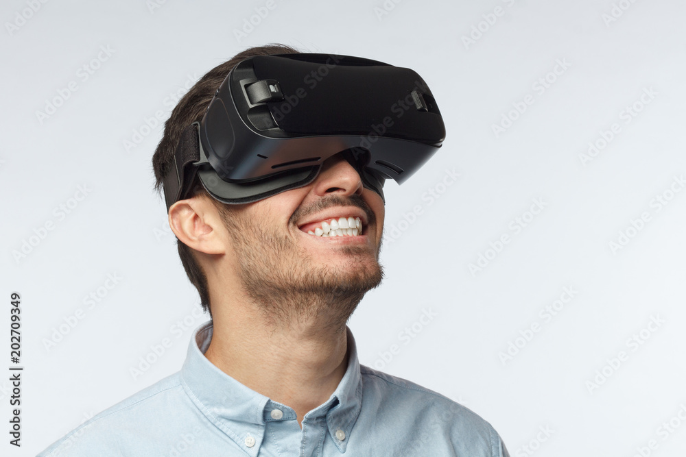 Horizontal closeup of young European Caucasian man with dark face hair isolated on grey background dressed casually, having VR headset on, experiencing its facilities with absolute satisfaction Horizontal closeup of young European Caucasian man with dark face hair isolated on grey background dressed casually, having VR headset on, experiencing its facilities with absolute satisfaction