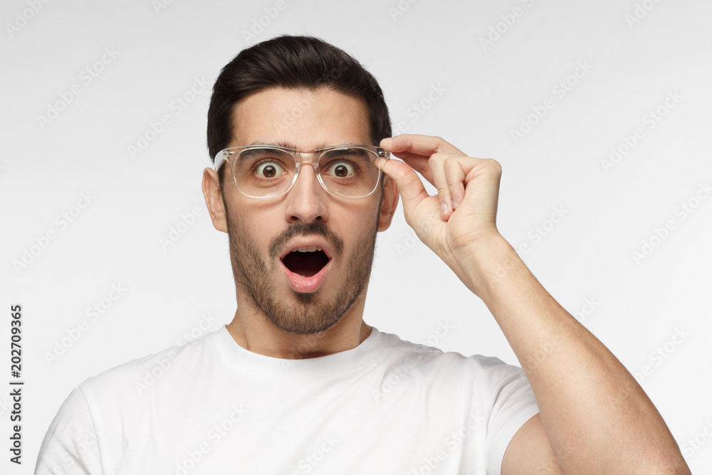 Studio portrait of young positive European Caucasian man isolated on grey background dressed in white T-shirt holding transparent plastic glasses looking with eyes and mouth round with surprise Studio portrait of young positive European Caucasian man isolated on grey background dressed in white T-shirt holding transparent plastic glasses looking with eyes and mouth round with surprise