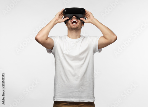 Horizontal half-length portrait of young European man pictured isolated on gray background wearing white T-shirt and virtual reality headset having lifted head up, laughing happily and having fun photo