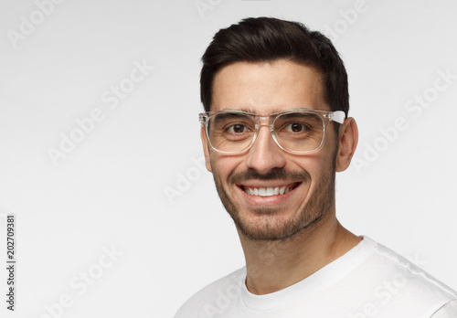 Indoor headshot of young handsome European man isolated on grey background in white casual T-shirt and with face hair, wearing trendy plastic eyeglasses and smiling friendly while looking at camera © Damir Khabirov