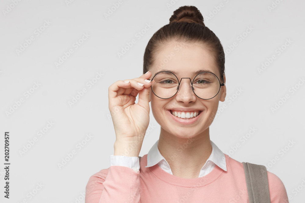 Closeup portrait of young European Caucasian woman pictured isolated on grey background, her hair tied in hairbun, holding rim of big round glasses with fingers, showing happy welcoming smile Closeup portrait of young European Caucasian woman pictured isolated on grey background, her hair tied in hairbun, holding rim of big round glasses with fingers, showing happy welcoming smile