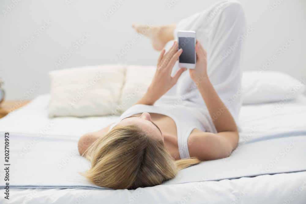 Blonde young woman using her smartphone while lying on her bed 