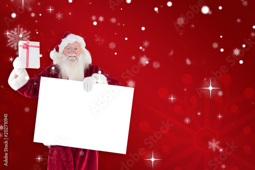 Santa shows a present while holding sign against red snowflake background © vectorfusionart