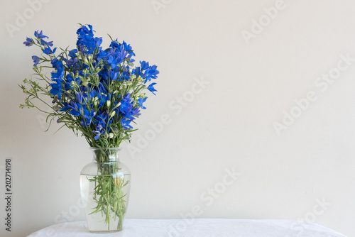 Delphiniums in glass vase on white table with tablecloth against neutral wall background (selective focus)