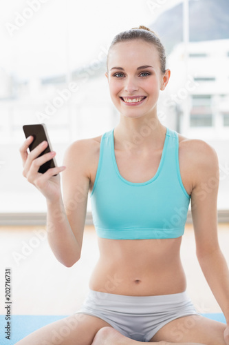 Sporty smiling woman holding phone while sitting on exercising mat