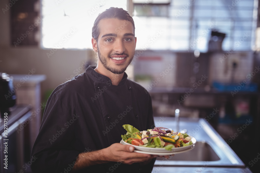 Portrait of smiling young waiter holding salad plate at commercial kitchen