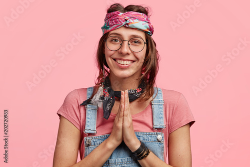 Obraz na plátně Positive young hippy woman with delighted look, keeps palms pressed together, as