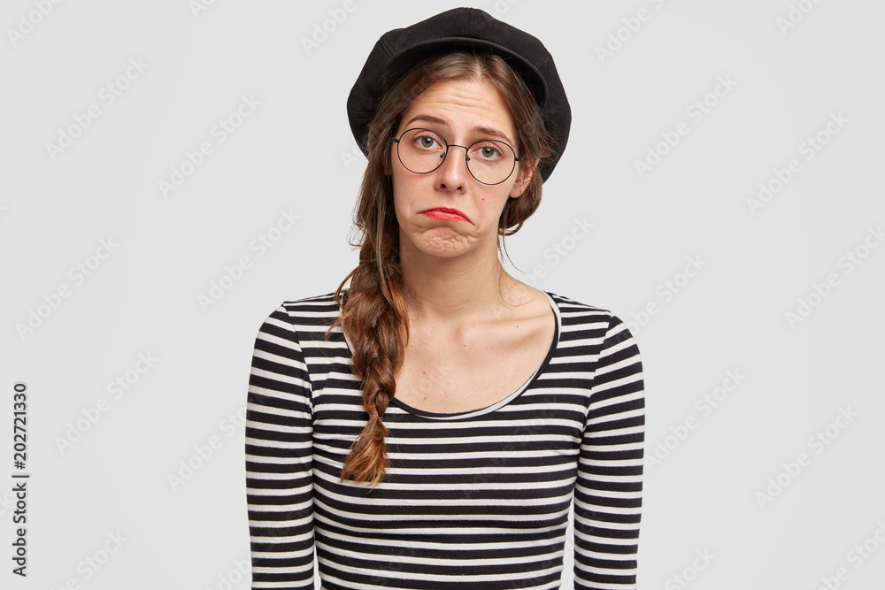 Discontent beautiful female with displeased expression, curves lips and looks with sad expression at camera, wears French style clothes, isolated over white studio background. Unhappy Parisian Discontent beautiful female with displeased expression, curves lips and looks with sad expression at camera, wears French style clothes, isolated over white studio background. Unhappy Parisian
