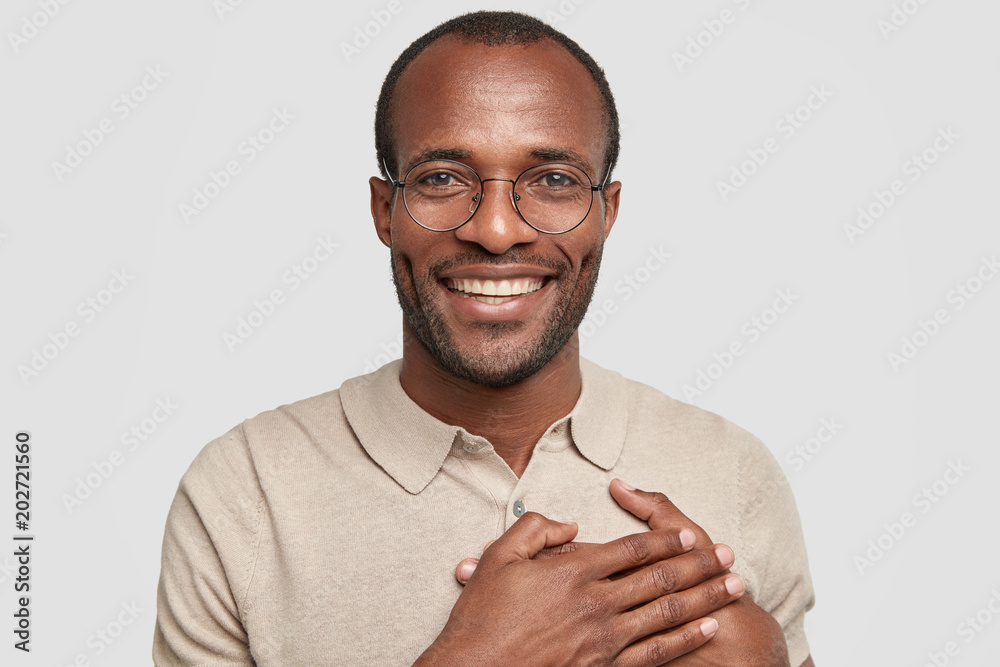 Φωτογραφία Dark skinned African American touched smiling male keeps hands on heart, looks joyfully at camera, has positive smile, expresses his friendly feelings to everybody, isolated over white background