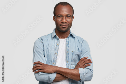 Image of confident attractive young African American male with pleased expression, keeps hands crossed, wears denim shirt and round spectacles, being content to recieve praisement from boss.