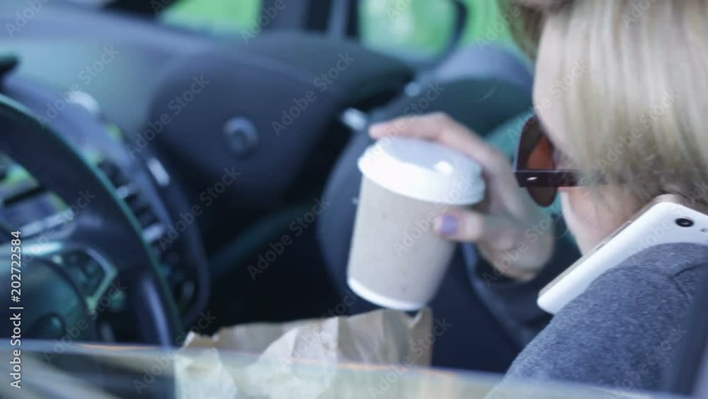 lady has an unhealthy lunch with burger and coffee inside car, talking on phone lady has an unhealthy lunch with burger and coffee inside car, talking on phone