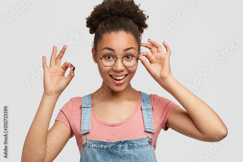 Curly beautiful dark skinned young woman with Afro hairstyle, demonstrates ok sign and shows her approval or like, has positive smile on face, wears fashionable clothes and round spectacles.
