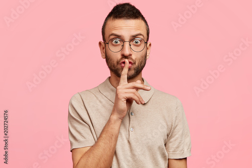 Shh, make silence and be quiet please! Surprised attractive young male with bristle, keeps fore finger on lips, shows hush or conspiracy sign, wears casual t shirt, isolated over pink background