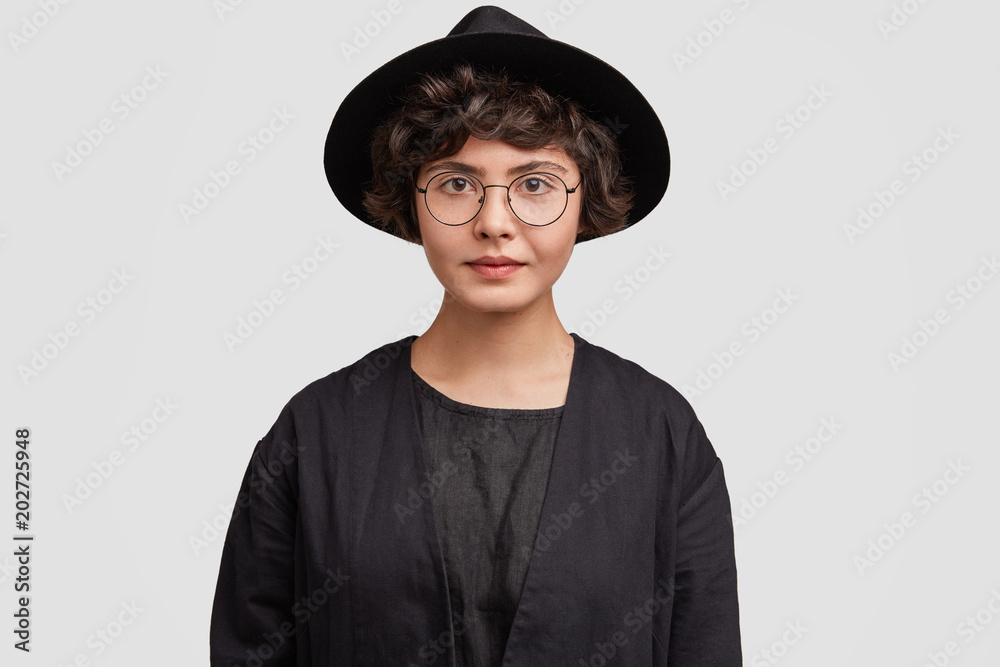 Portrait of serious mixed race female student with curly hair, wears black clothes and round spectacles, looks confidently at camera, isolated over white background. Lovely gorgeous woman indoor Portrait of serious mixed race female student with curly hair, wears black clothes and round spectacles, looks confidently at camera, isolated over white background. Lovely gorgeous woman indoor