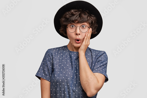 Candid shot of stunned hipster woman looks with great surprisement at camera, keeps mouth opened, wears fashionble polka dot blouse and big black hat, expresses shock, isolated on white background photo