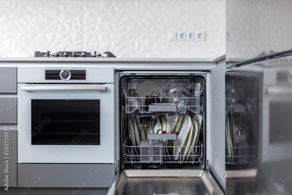 Opened dishwasher machine with dishes inside just washed, in a kitchen with grey fronts, white backsplash and white quartz countertop. Opened dishwasher machine with dishes inside just washed, in a kitchen with grey fronts, white backsplash and white quartz countertop.