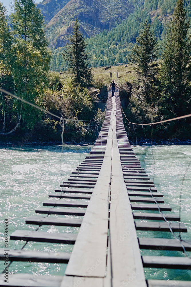 Young man walks along a suspension bridge across a mountain river Katun in the early autumn