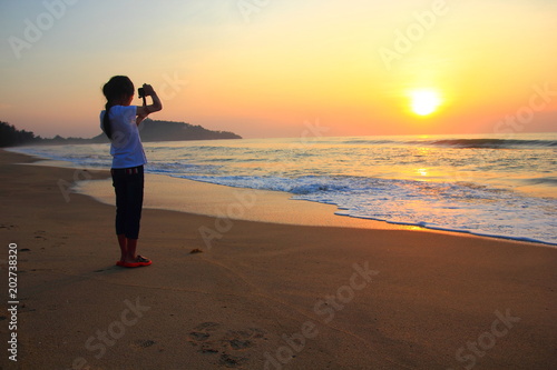 Little girl taking photos on the beach Kid girl on the beach with phone taking photos of a seascape view. child is making selfie on a sunset ,Thailand