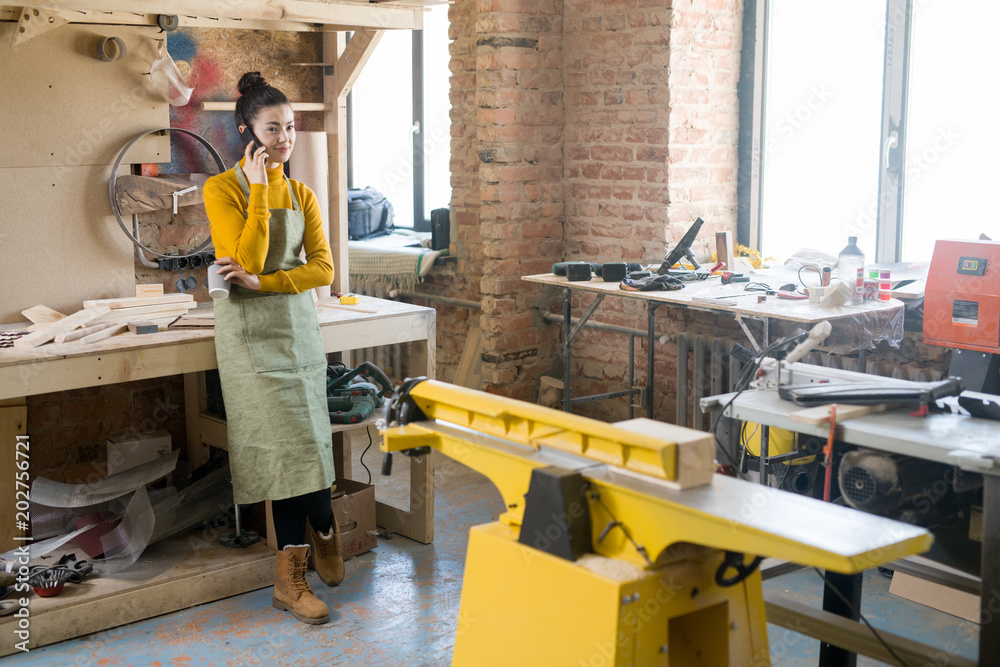 Full length portrait of modern young woman speaking by phone while working in loft-styled carpenters manufactory, copy space Full length portrait of modern young woman speaking by phone while working in loft-styled carpenters manufactory, copy space