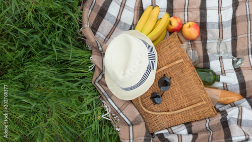 Plaid, hat, glasses, book, wine with a basket on a plaid on the green grass. The concept of a picnic, summer and rest. Top view with empty space for labeling or advertising