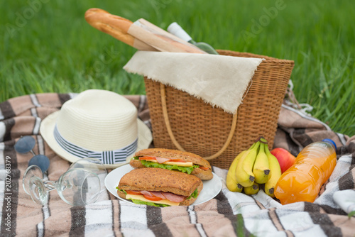 Plaid, hat, glasses, book, senvichi, juice and fruit with a basket on a plaid on the green grass. The concept of a picnic, summer and rest. Top view with empty space for labeling or advertising
