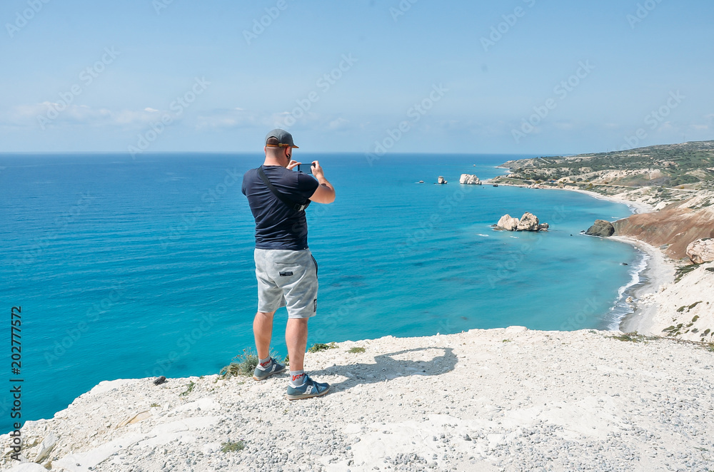 Man near sea making photo with his mobile phone camera.Young man standing on top of cliff in Cyprus, Mediterranean Sea. Travel, vacation concept. Copyplace, place for text