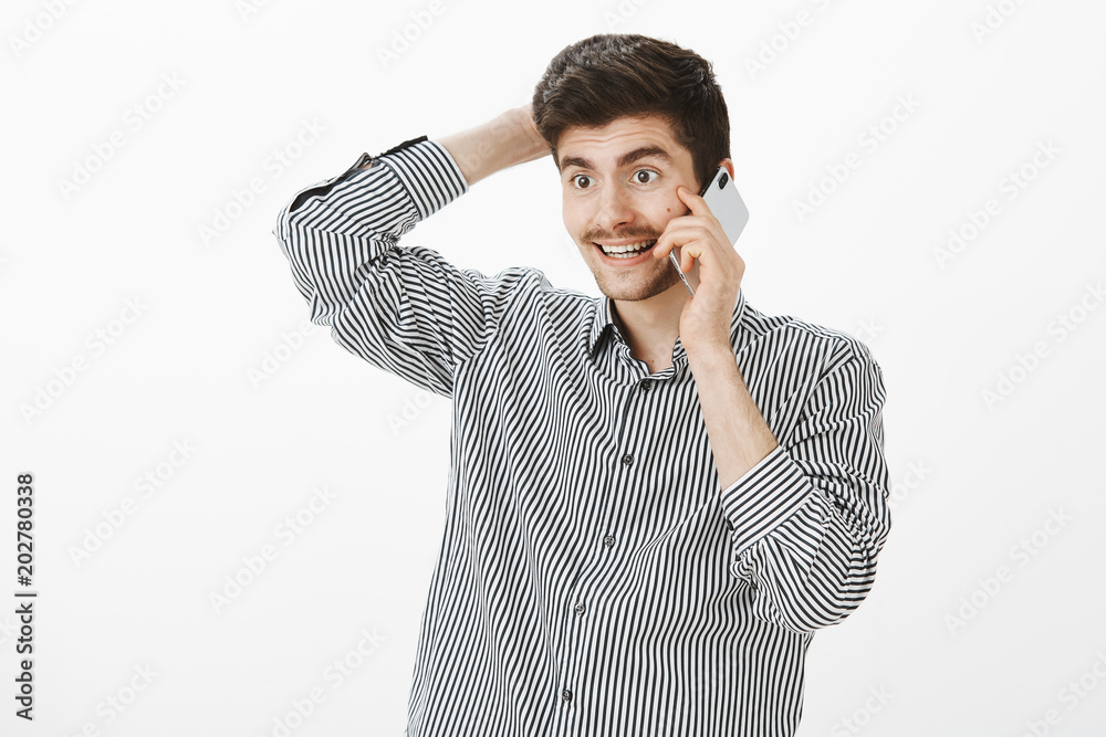Guy surprised with call. Portrait of pleased joyful young european bearded man, scratching head and talking on smartphone, smiling, feeling awkward and shy, standing over gray background Guy surprised with call. Portrait of pleased joyful young european bearded man, scratching head and talking on smartphone, smiling, feeling awkward and shy, standing over gray background