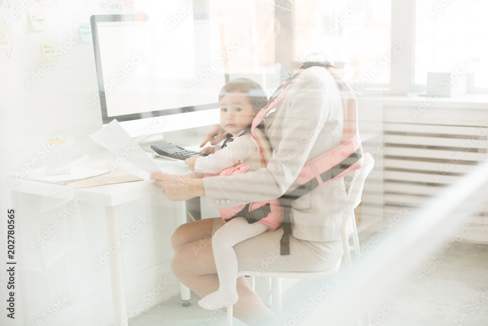 Baby daughter of young accountant sitting in front carrier close to her mother during work in office Baby daughter of young accountant sitting in front carrier close to her mother during work in office