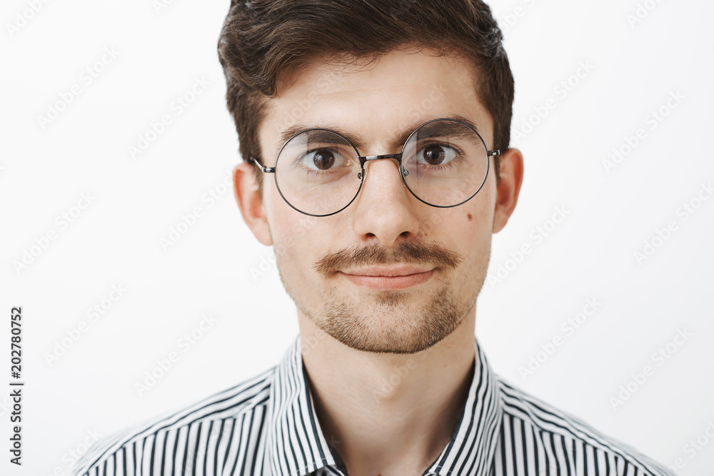 Close-up shot of confident smart creative guy with beard and moustache in round glasses, standing carefree and self-assured over gray background, talking casually or hearing out friend over gray wall