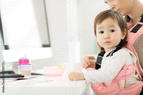 Cute baby girl in pink carrier and her business mother sitting by desk in office during lunch break