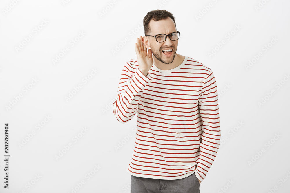 What did you say, repeat. Portrait of annoyed questioned attractive european guy in black glasses, asking question and holding hand near ear, hearing nothing while standing in loud place What did you say, repeat. Portrait of annoyed questioned attractive european guy in black glasses, asking question and holding hand near ear, hearing nothing while standing in loud place