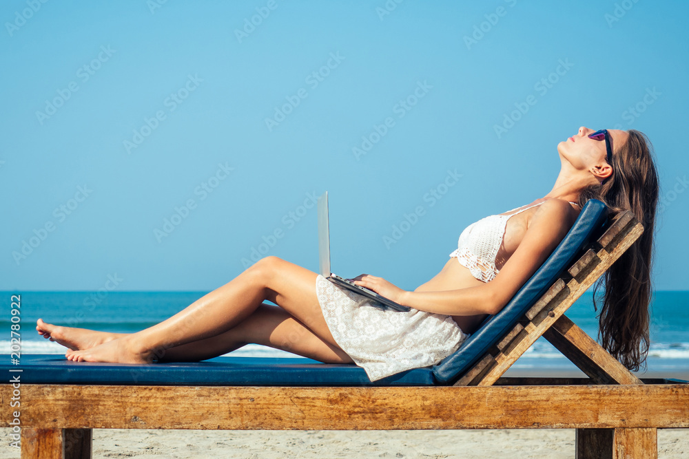 beautiful and sexy young woman is sitting on a deckchair and is resting with a laptop on her knees on the sea. woman freelancer is resting with laptop in the resort