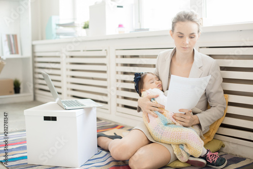 Young economist reading paper while sitting on office floor with her sleeping baby daughter