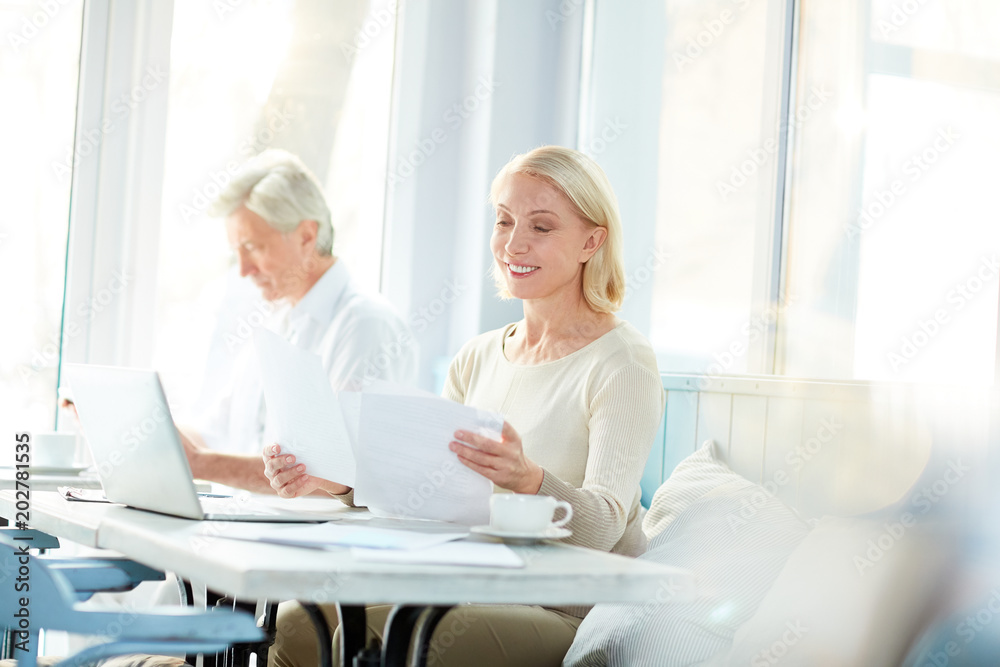 Two contemporary senior accountants sitting by table in comfortable cafe, working with papers and browsing in the net Two contemporary senior accountants sitting by table in comfortable cafe, working with papers and browsing in the net
