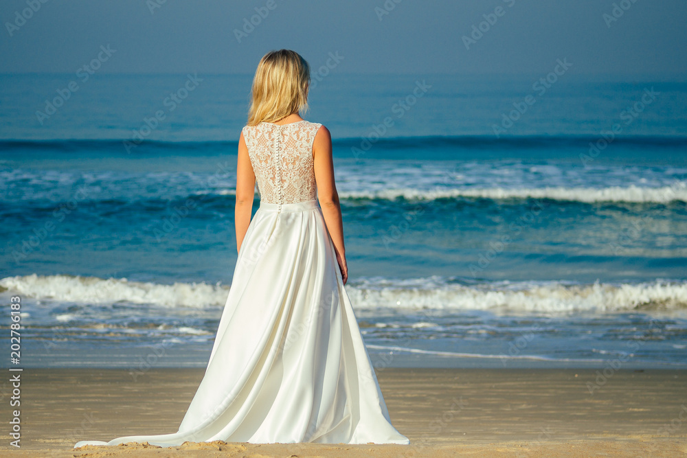 young and beautiful bride with long blond hair standing his back and looking at the sea in a beautiful white long wedding dress. a happy bride married a wedding ceremony on the beach by the sea young and beautiful bride with long blond hair standing his back and looking at the sea in a beautiful white long wedding dress. a happy bride married a wedding ceremony on the beach by the sea