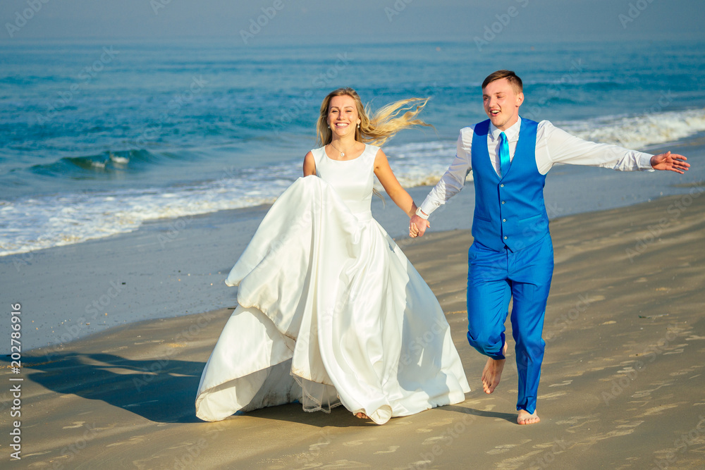 handsome groom in a chic suit and a beautiful bride in a wedding dress are having fun and running on the beach. concept of a chic and rich wedding ceremony on the beach