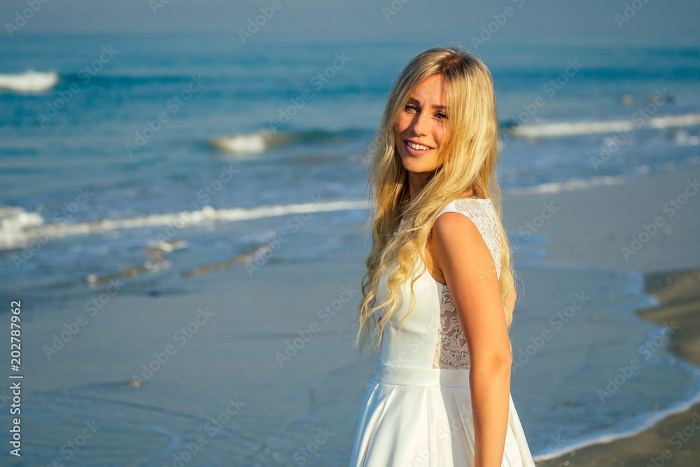 young and beautiful bride with long blond hair beautifully smiling and walking on the sea in beautiful white long wedding dress. a happy bride married a wedding ceremony on the beach by the sea
