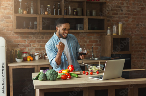 Happy black man looking at laptop and cooking dinner