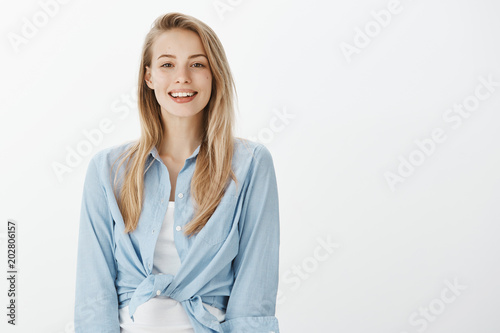 Portrait of kind charming european female student in stylish outfit, smiling friendly and happilly while gazing at camera, expressing positive, joyful attitude while talking casually over white wall