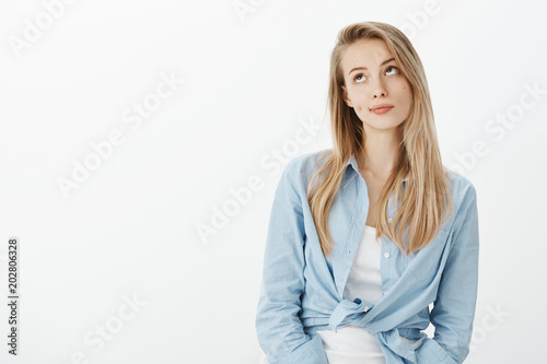 Portrait of unimpressed beautiful european woman with blond hair, smirking and looking upwards with indifferent expression, standing in stylish outfit over white background casually