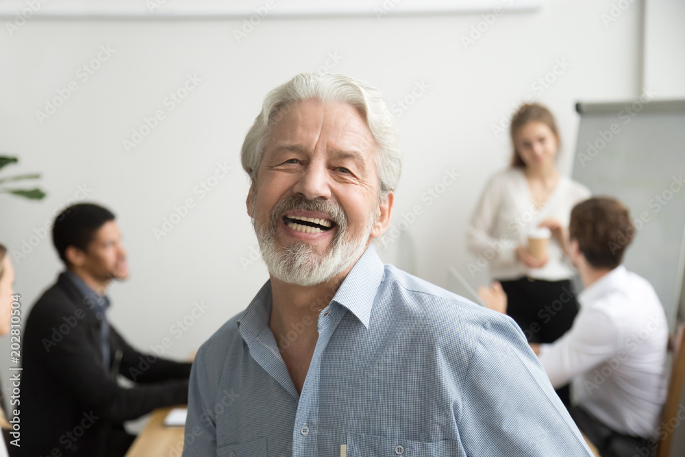 Happy senior businessman laughing looking at camera in office, cheerful team leader posing with employees at background, aged teacher, old gray-haired boss or professional mentor head shot portrait Happy senior businessman laughing looking at camera in office, cheerful team leader posing with employees at background, aged teacher, old gray-haired boss or professional mentor head shot portrait