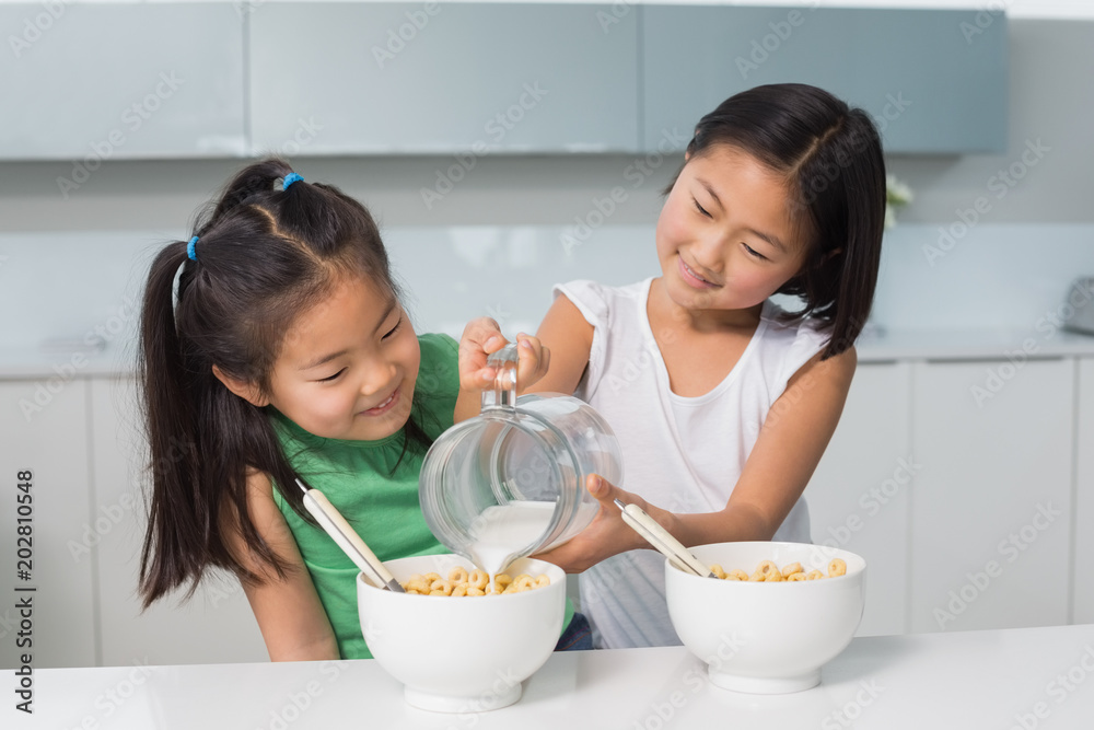Two happy young girls pouring milk in bowl in kitchen Two happy young girls pouring milk in bowl in kitchen
