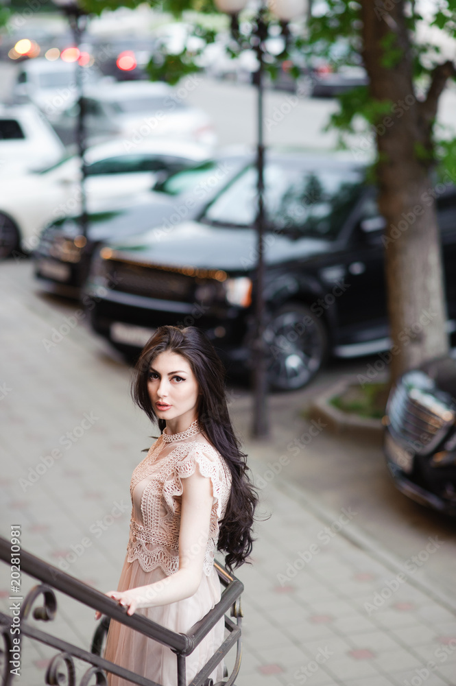 fashionable pretty girl in summer dress on background of city streets with expensive cars and green trees outdoor