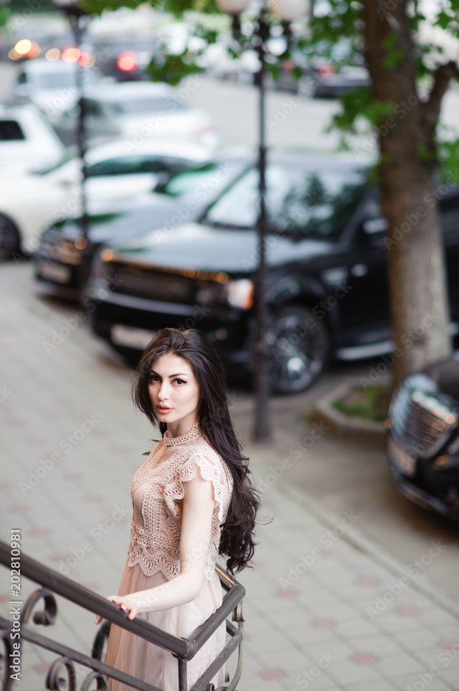 Fototapeta premium fashionable pretty girl in summer dress on background of city streets with expensive cars and green trees outdoor