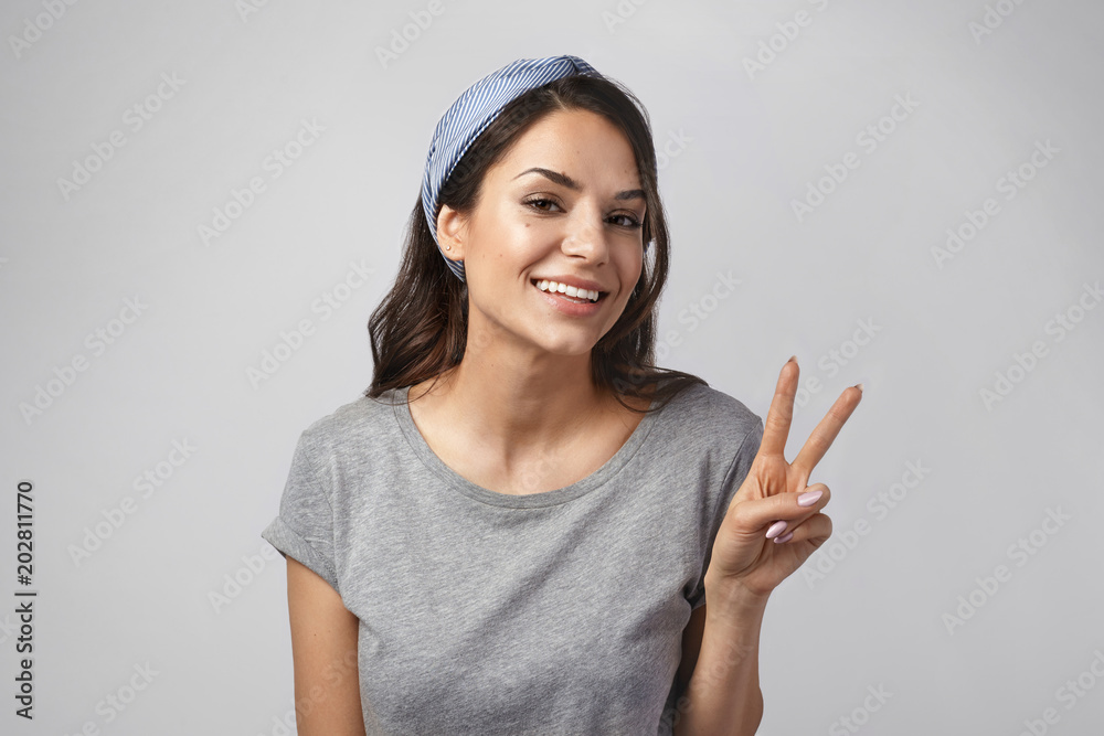 Positive human emotions and body language. Portrait of attractive fashionable dark haired female posing in studio, smiling broadly and making peace gesture, her look expressing friendliness Positive human emotions and body language. Portrait of attractive fashionable dark haired female posing in studio, smiling broadly and making peace gesture, her look expressing friendliness