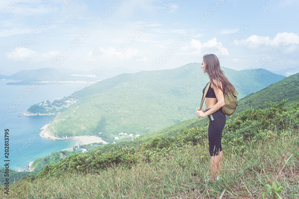 Naklejka premium Backpack female traveler standing on hill looking at sea and mountains. Trail runner taking a break enjoying view of nature