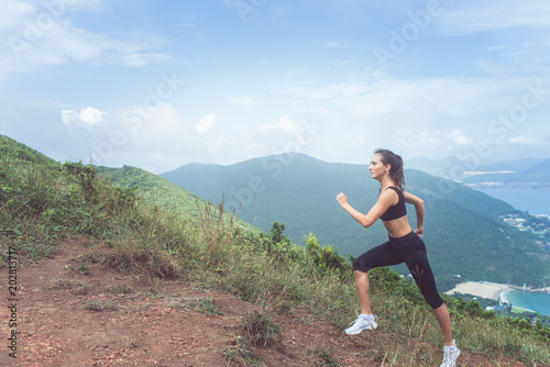 Slender young female athlete doing cardio exercise going up the mountain with sea in background.